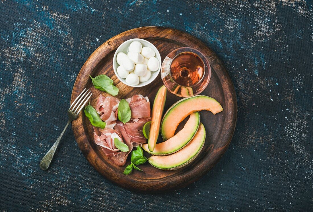 Slices of cantaloupe melon, Parma ham, fresh basil leaves, mozzarella and a glass of rosé wine on a wooden board