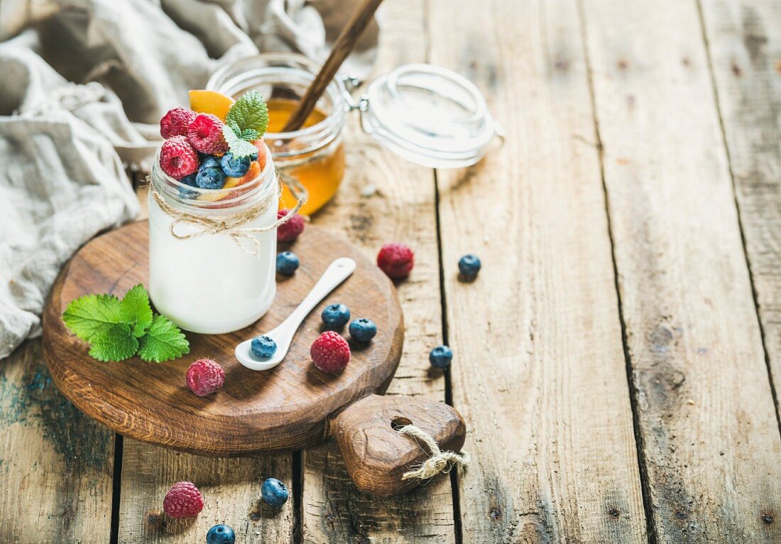 A glass jar of white yoghurt with fresh berries, peach and mint on a wooden serving board