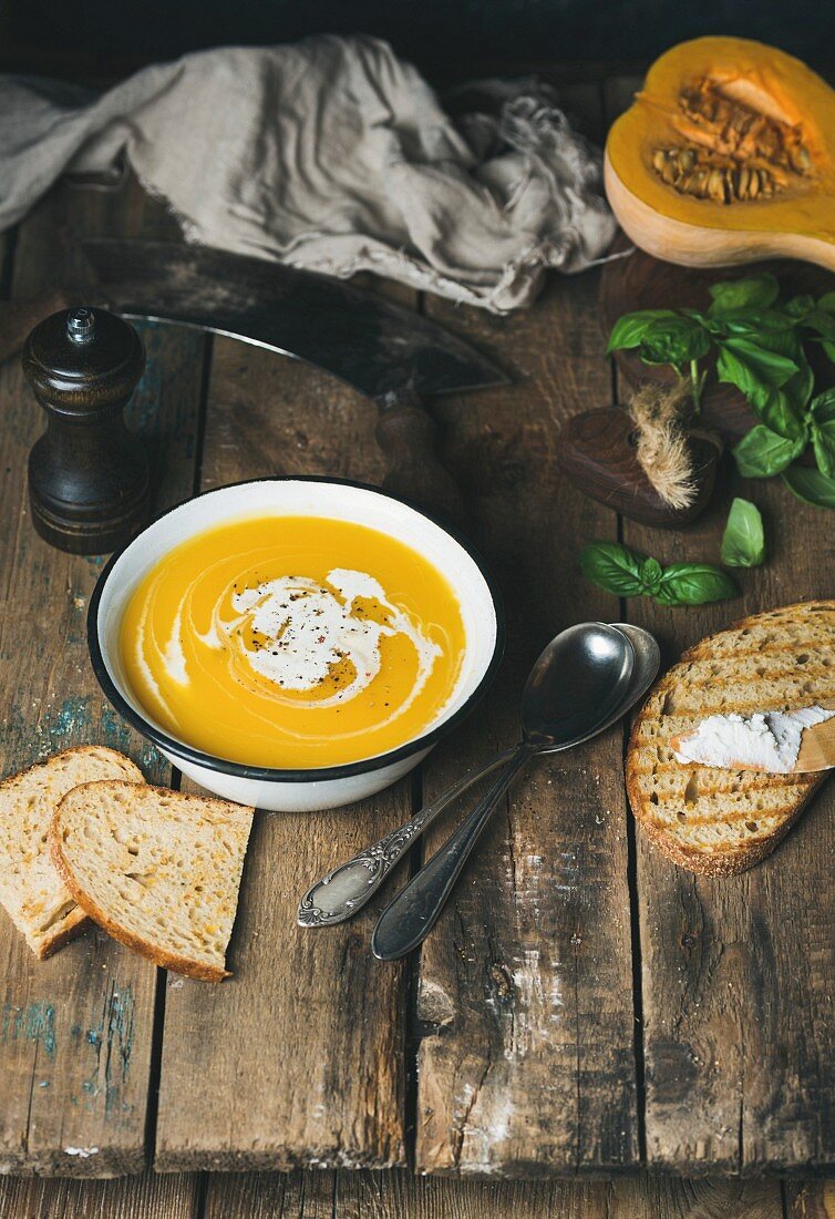 Creamy pumpkin soup in a white bowl with fresh basil and grilled bread slices on a rustic wooden table