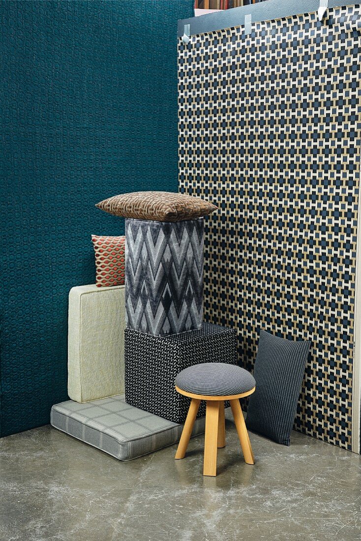 A pile of cushions, a stool and seating blocks in front of two walls with patterned wallpapers