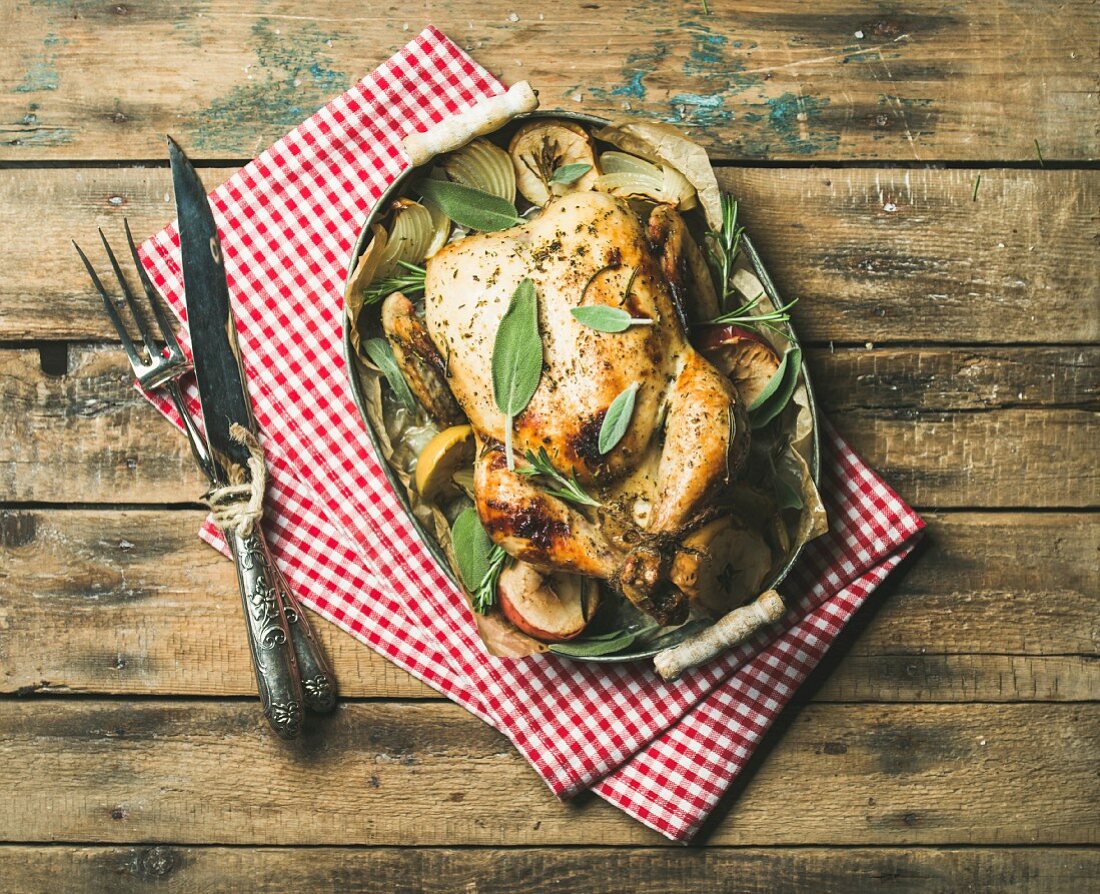 Oven roasted whole chicken with onion, apples and sage in serving tray over rustic wooden background