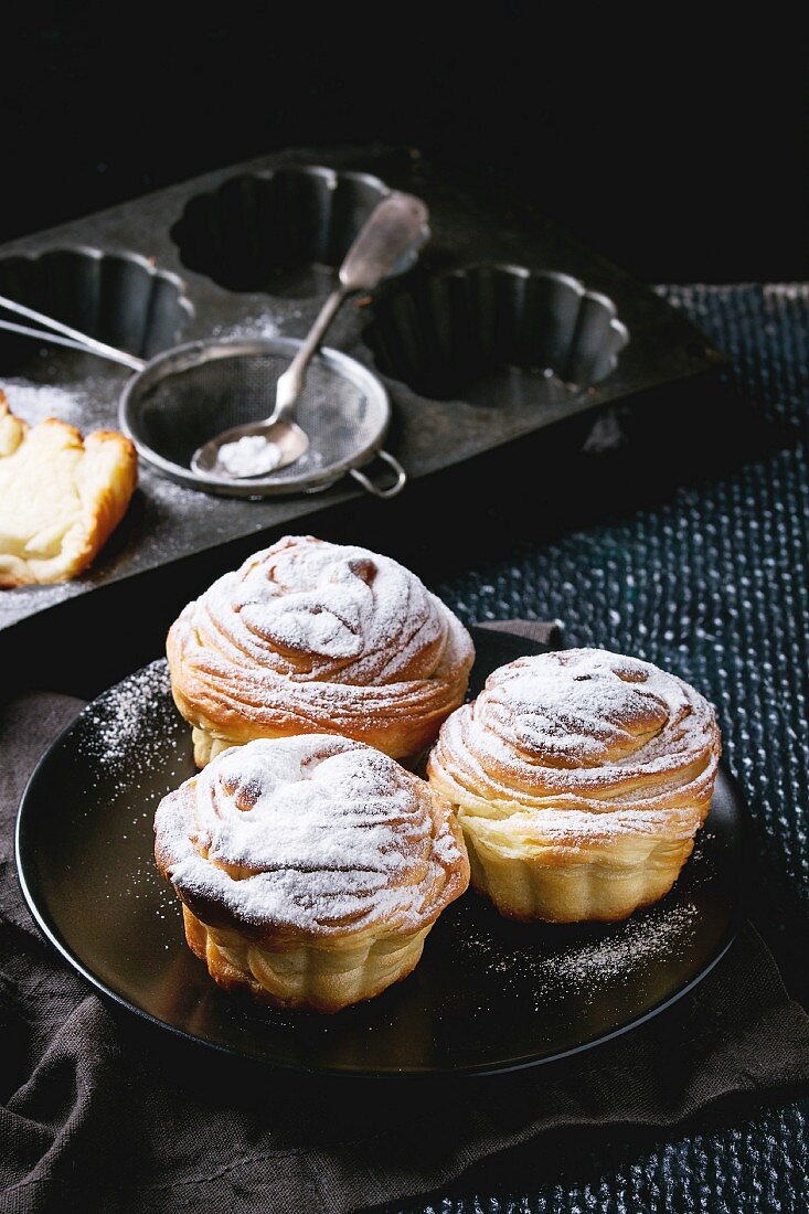 Modern pastries cruffins, whole and slice, with sugar powder served on black plate with sieve