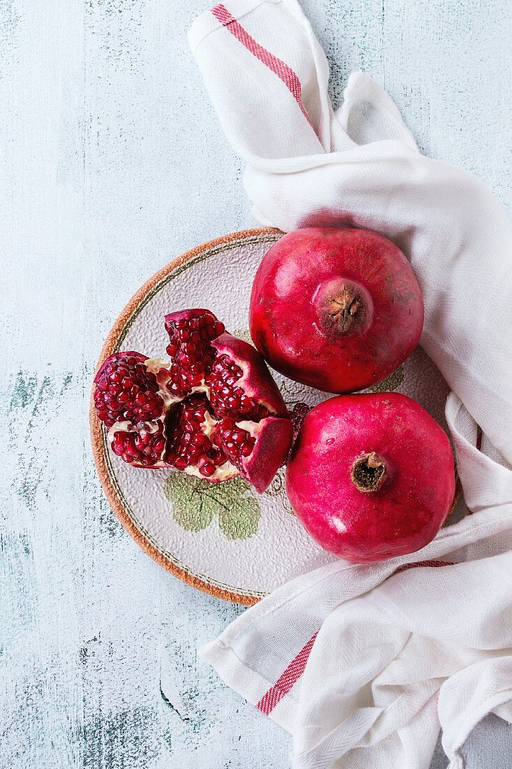 Ripe slice and whole pomegranates on ornate ceramic plate on kitchen linen towel over white wooden background