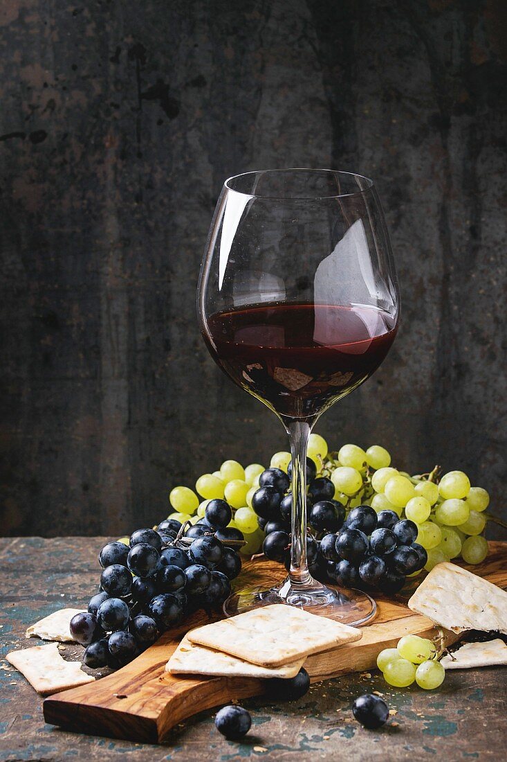 Bunches of ripe wet red and white grapes with crackers snack and glass of red wine on olive wood cutting board