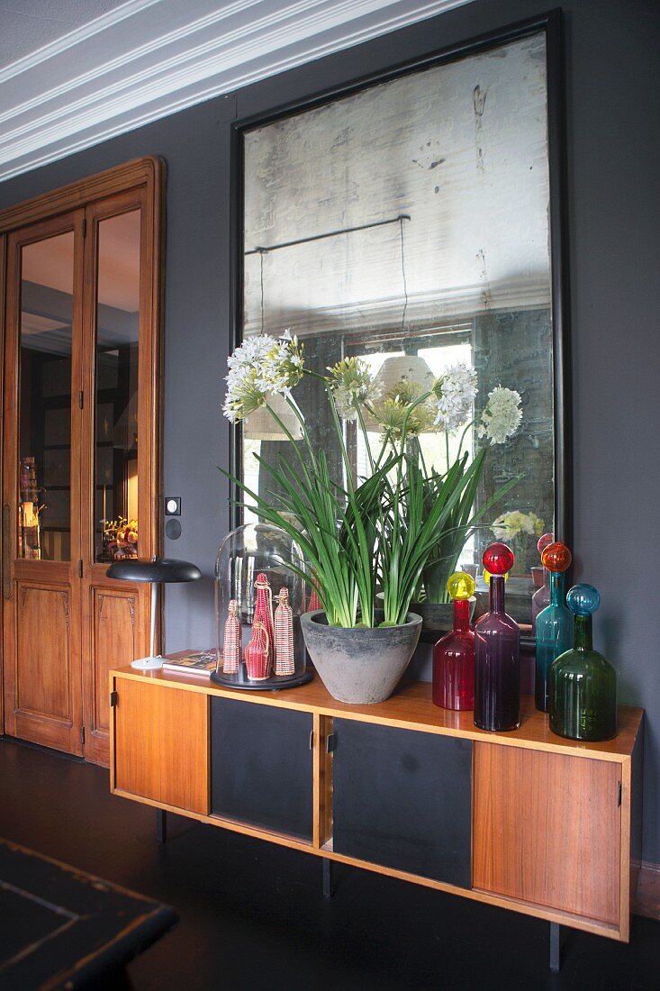 Retro Sideboard mit bunten Glasflaschen und Pflanzschale mit weißen Blüten vor Wandspiegel
