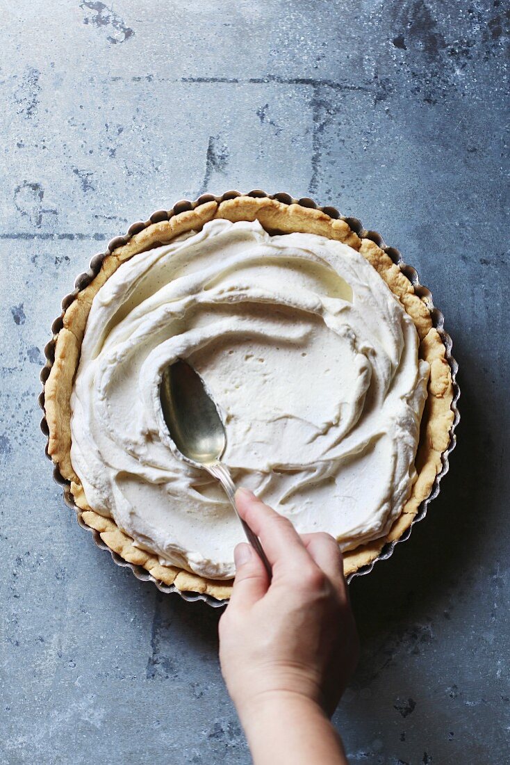 Hand spreading with the back of a spoon the whipped cream filling over the bottom of a tart