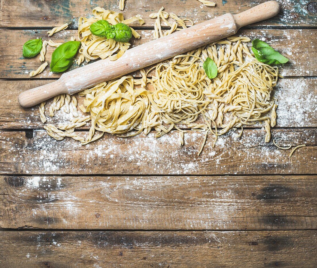 Various homemade fresh uncooked Italian pasta with flour, green basil leaves and plunger on shabby background