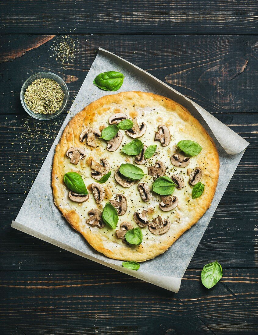 Pizza mit Pilzen und Basilikum auf Backpapier, Gewürzmischung im Glas