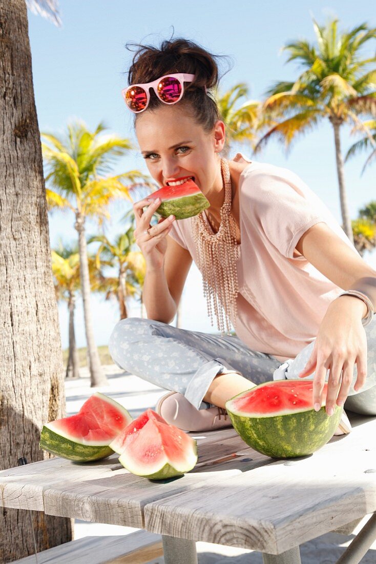 Brünette Frau in cremefarbenem T-Shirt und hellgrauer Hose isst am Strand eine Melone