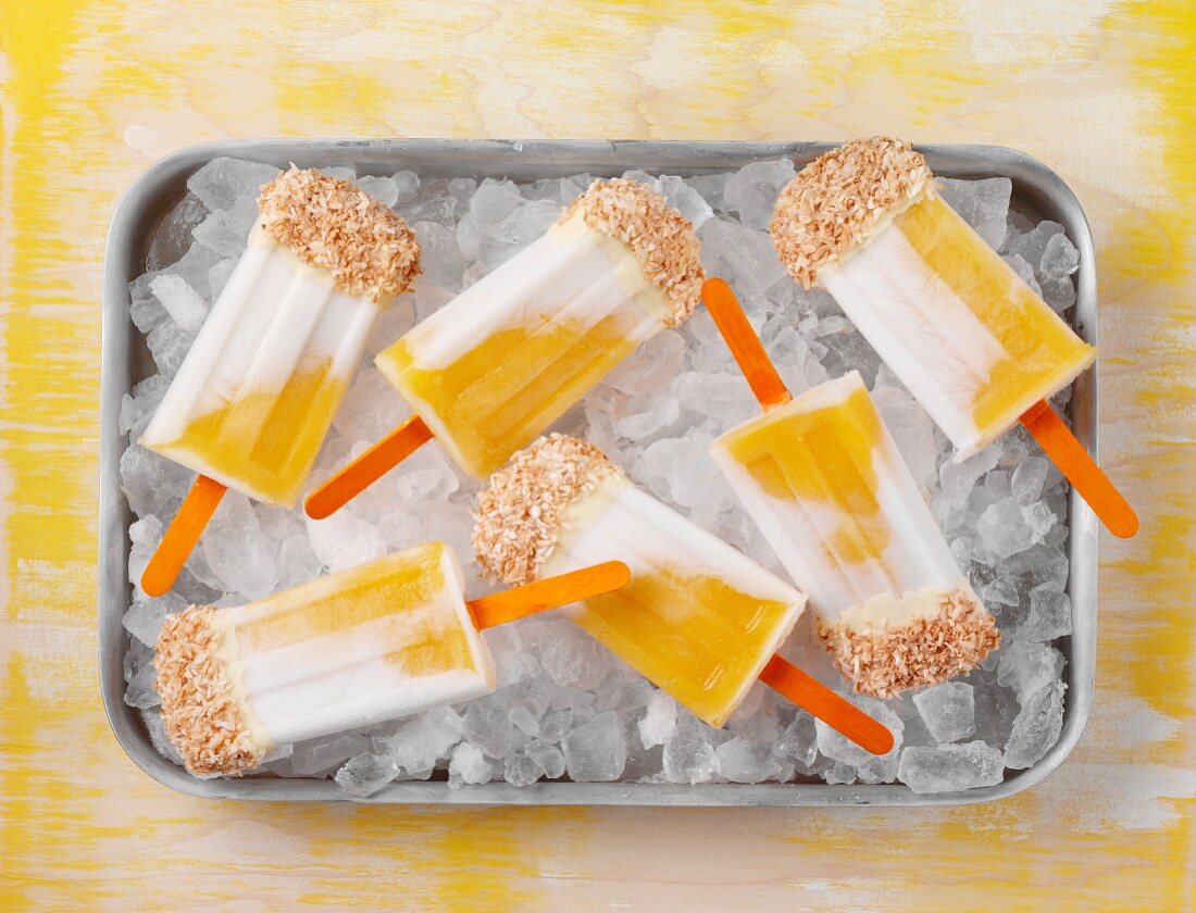 Mango and coconut ice lollies topped with white chocolate and desiccated coconut on a bed of ice cubes