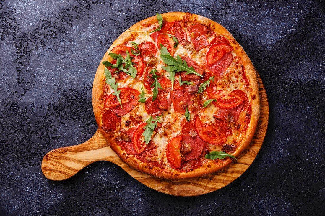 Pizza with ham, tomato and arugula leaves on wood olive cutting board on dark background