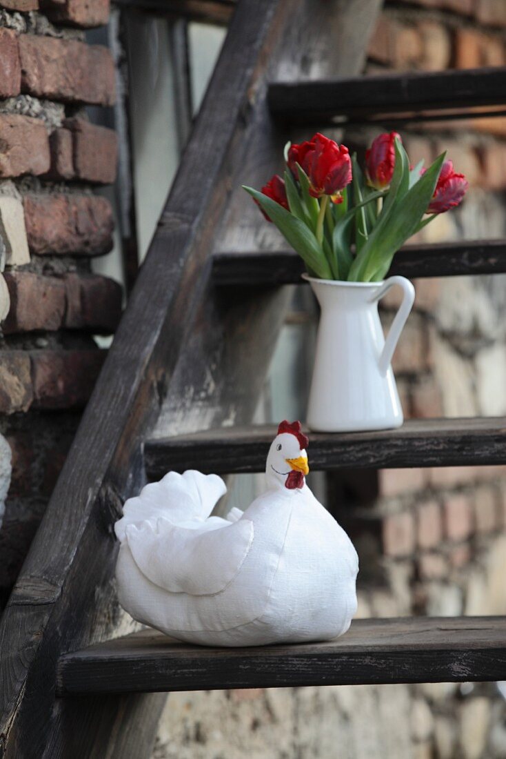 Hand-made fabric hen and white jug of red tulips on rustic wooden steps