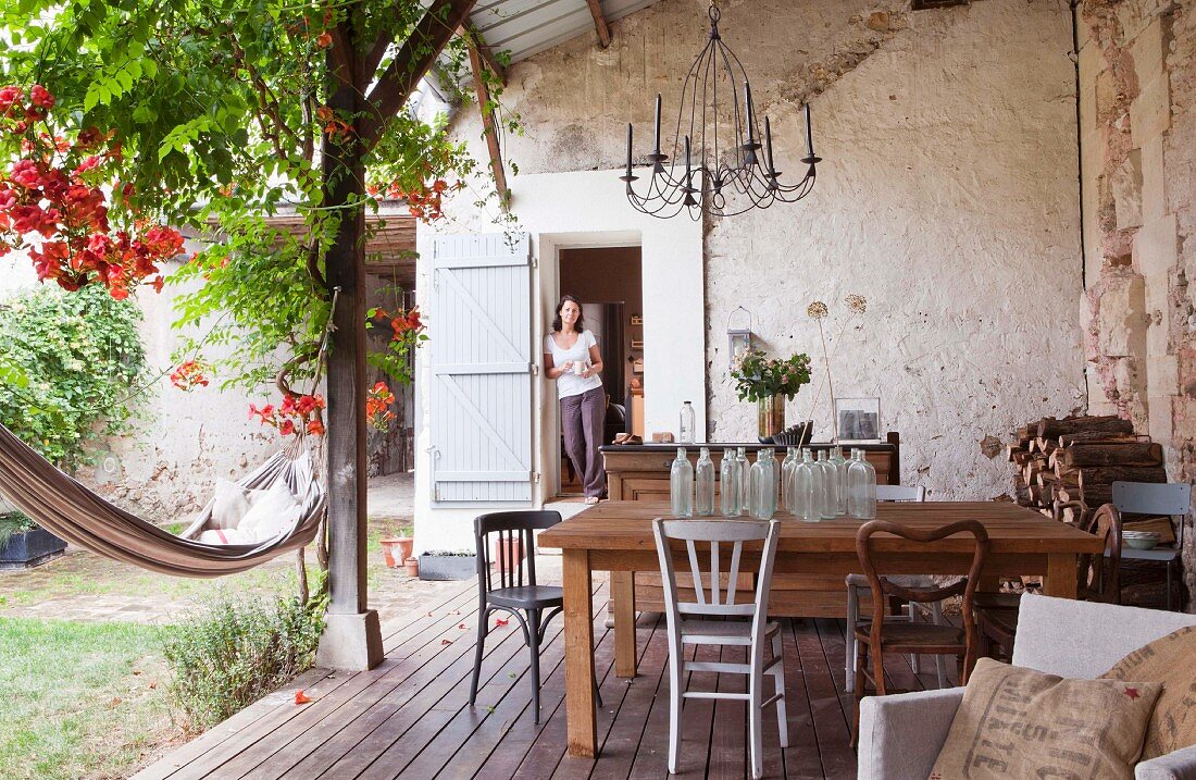 Wooden table with collection of glass bottles on roofed country-house terrace; woman in doorway in background