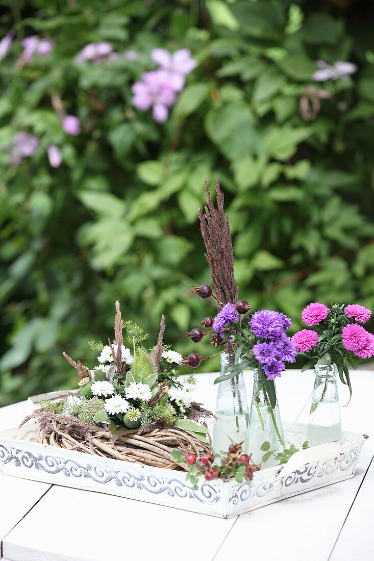 Wreath decorated with white asters, thistles and poppy seedheads next to pink asters in glass bottles