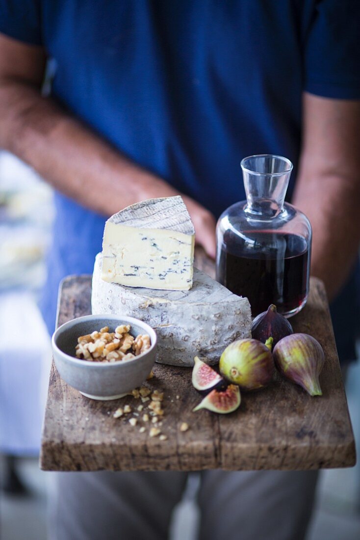 A man holding a cheese board with blue cheese, figs, nuts and wine