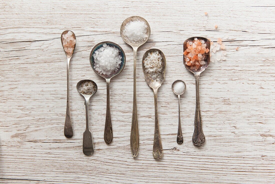 Seven vintage spoons of diffrent types of salt on a white wooden surface