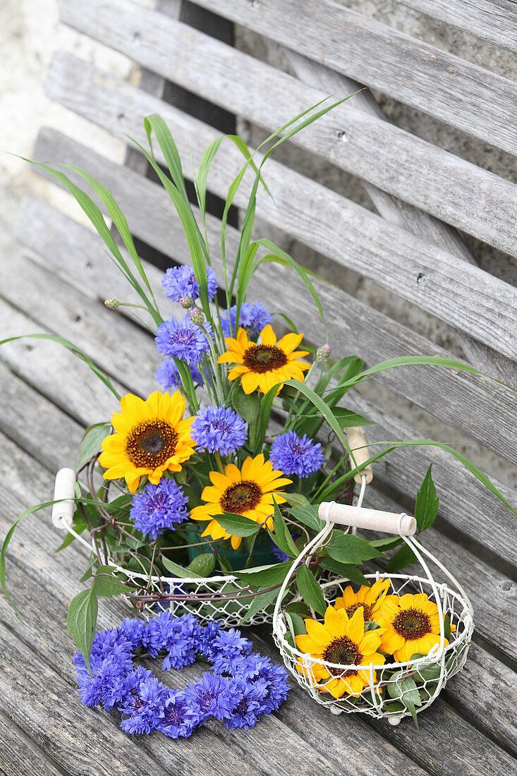 Sunflowers, cornflowers, blades of grass and clematis tendrils in wire basket next to wreath of cornflowers on wooden bench