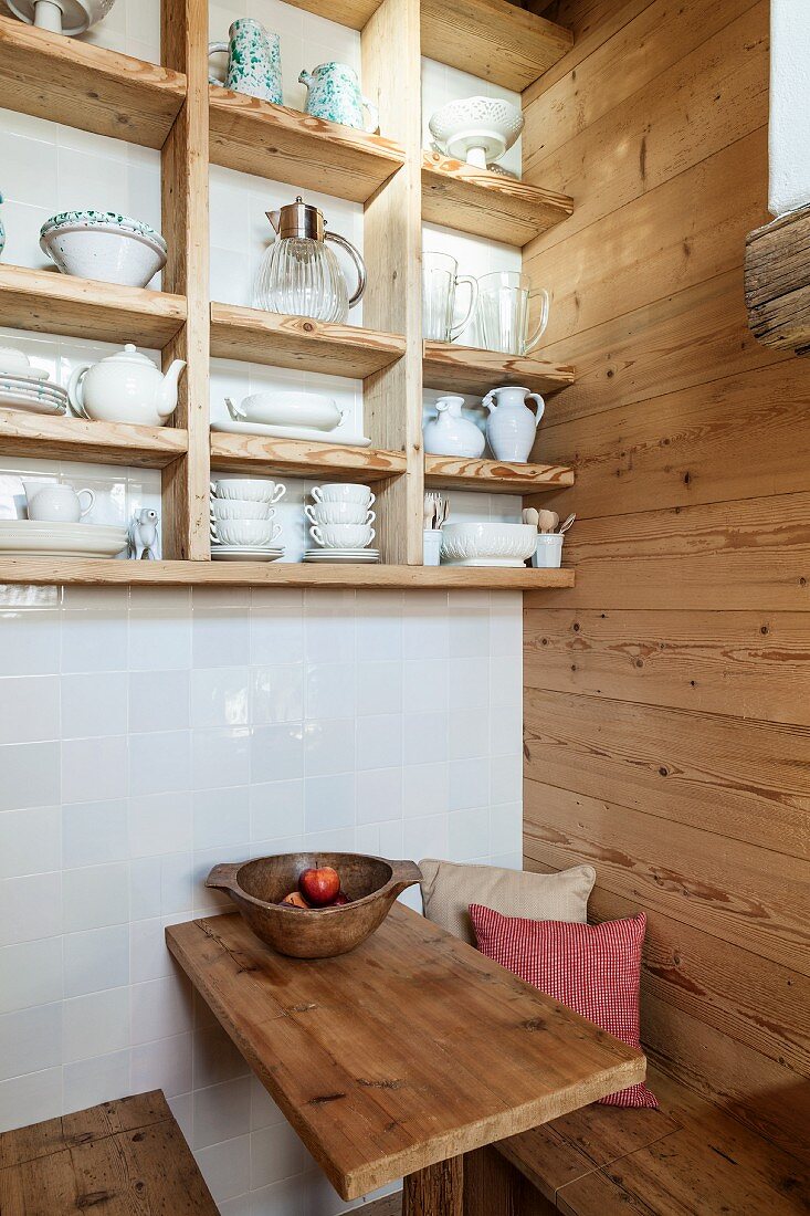 Crockery on open-fronted shelves on white-tiled wall above wooden benches and wooden table in seating area