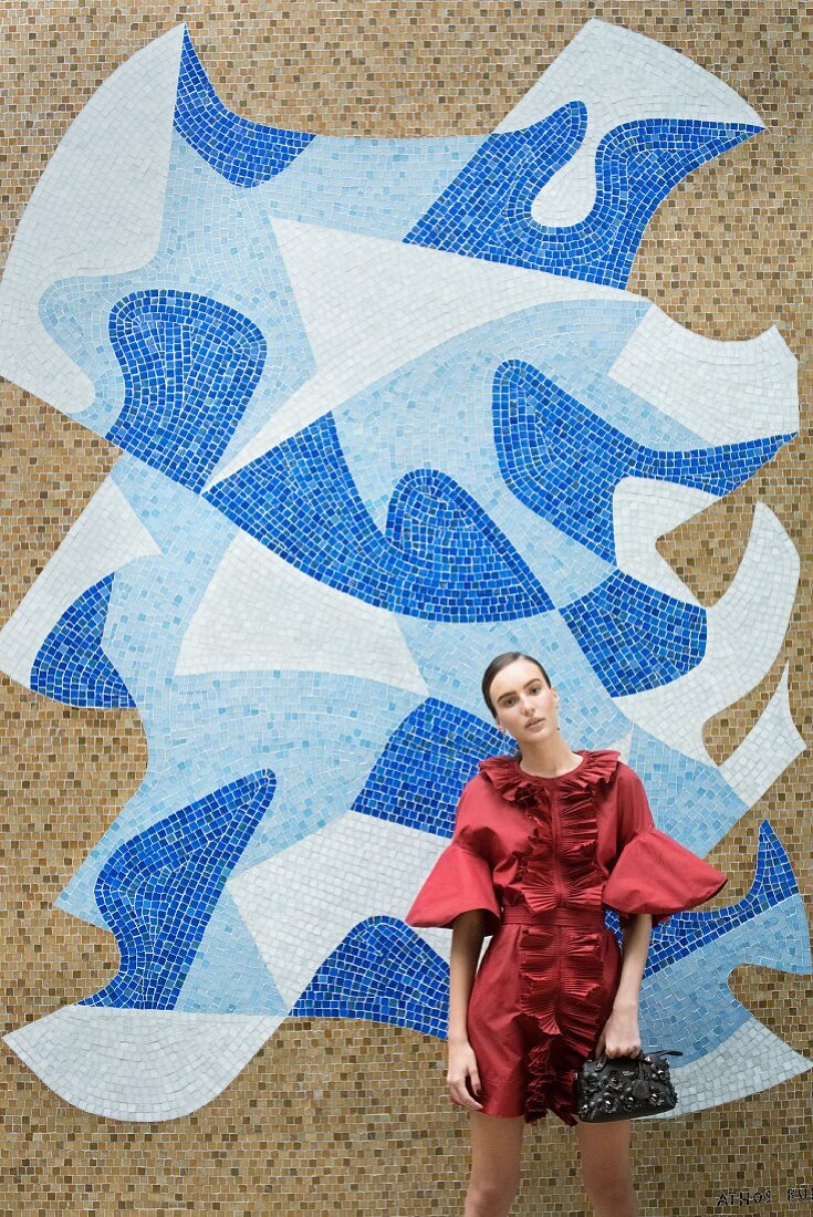 A brunette woman wearing a red ruched dress and holding a black handbag in front of a mosaic wall