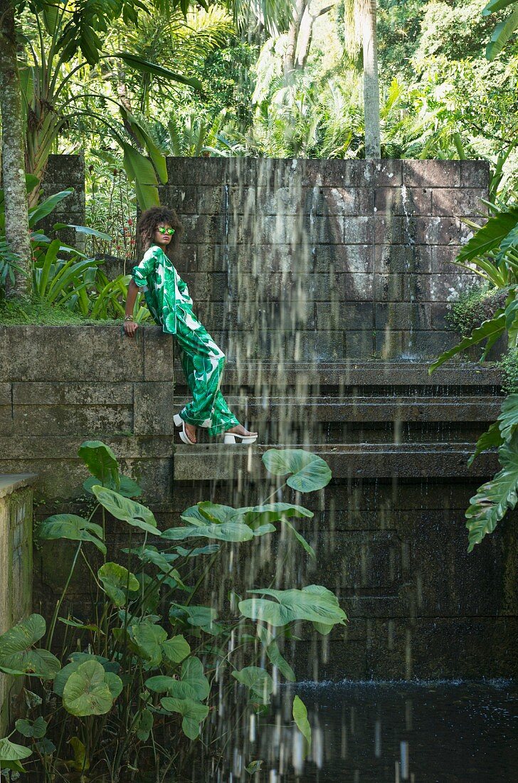 Brünette Frau in Hosenanzug mit tropischem Print, Sandalen und Sonnenbrille hinter einem Wasserfall