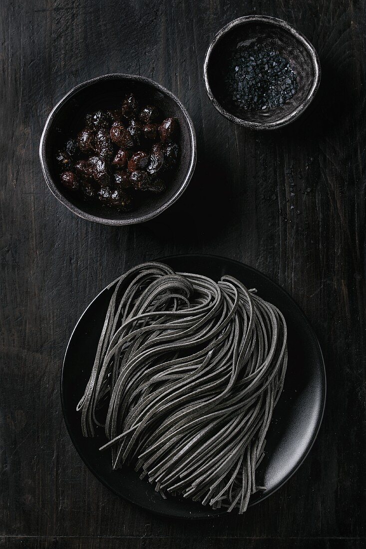 Ungekochte schwarze Tintenfischspaghetti mit schwarzen Oliven und schwarzem Salz auf schwarzem Hintergrund