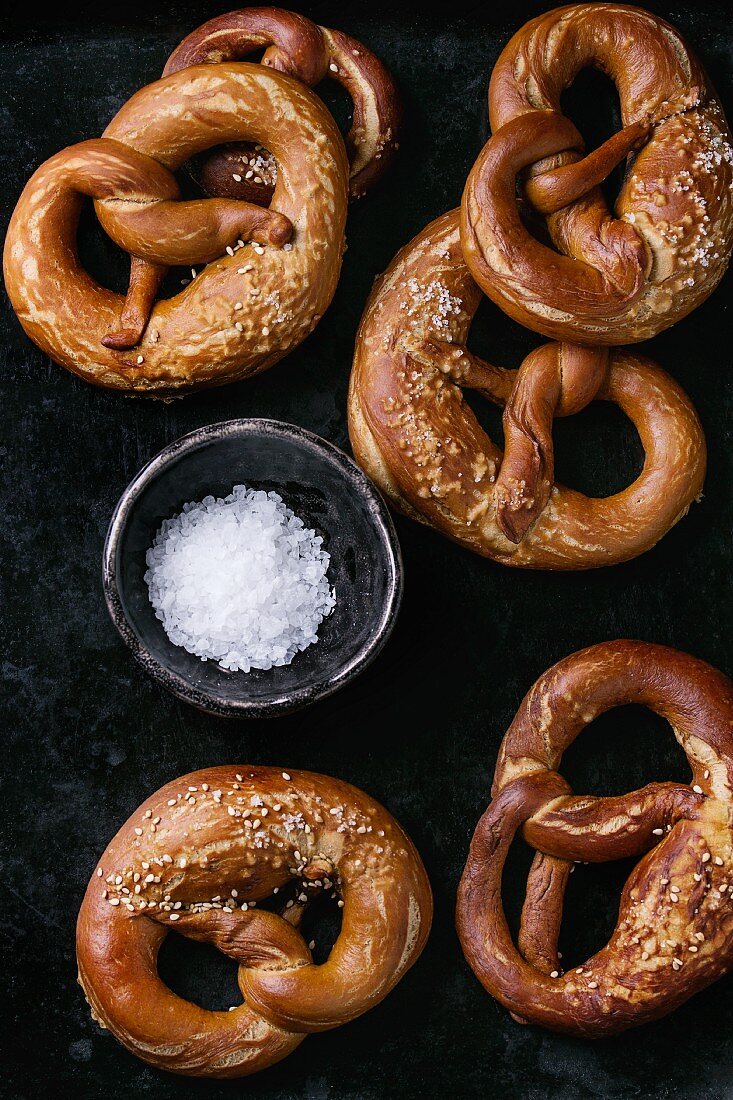 Traditional salted pretzels and bowl of sea salt over black background View from above