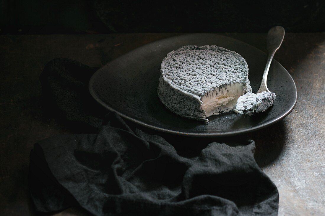 Goat cheese with gray mold with fork on dark plate over black background