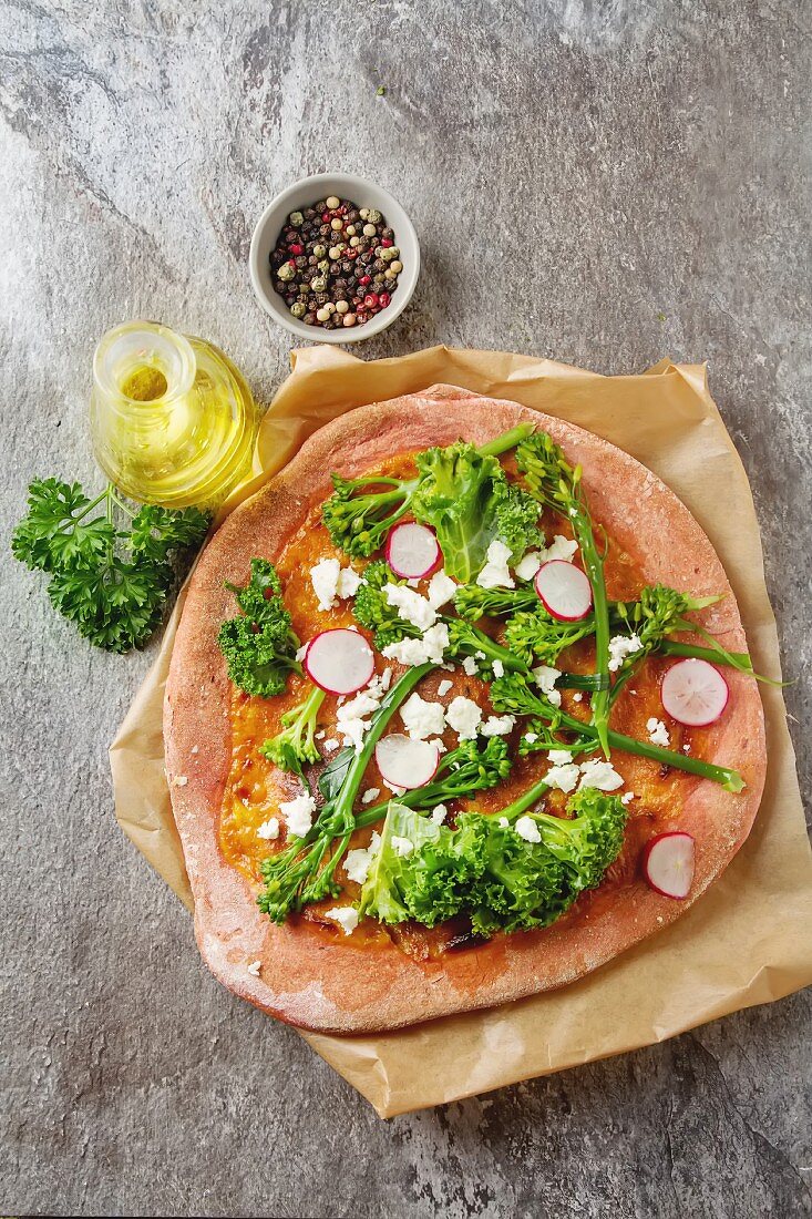 Pizza aus Rote-Bete-Teig mit Brokkoli, Salat, Radieschen und Feta, daneben Olivenöl, Pfeffer und Petersilie