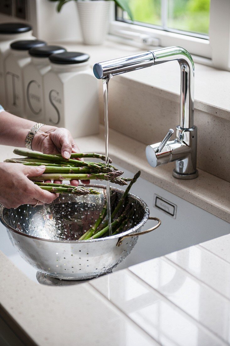 Hands washing green asparagus in colander under running water