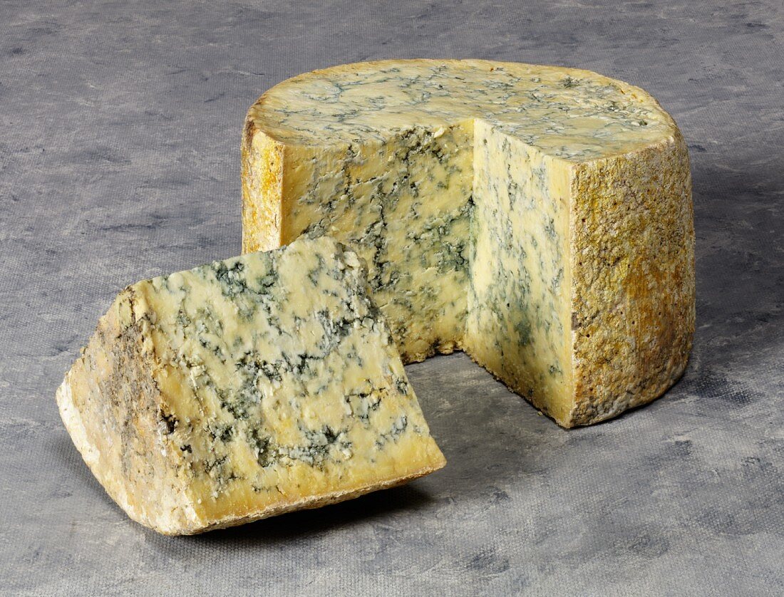 A whole stilton cheese with a quarter cut out shown on a grey background