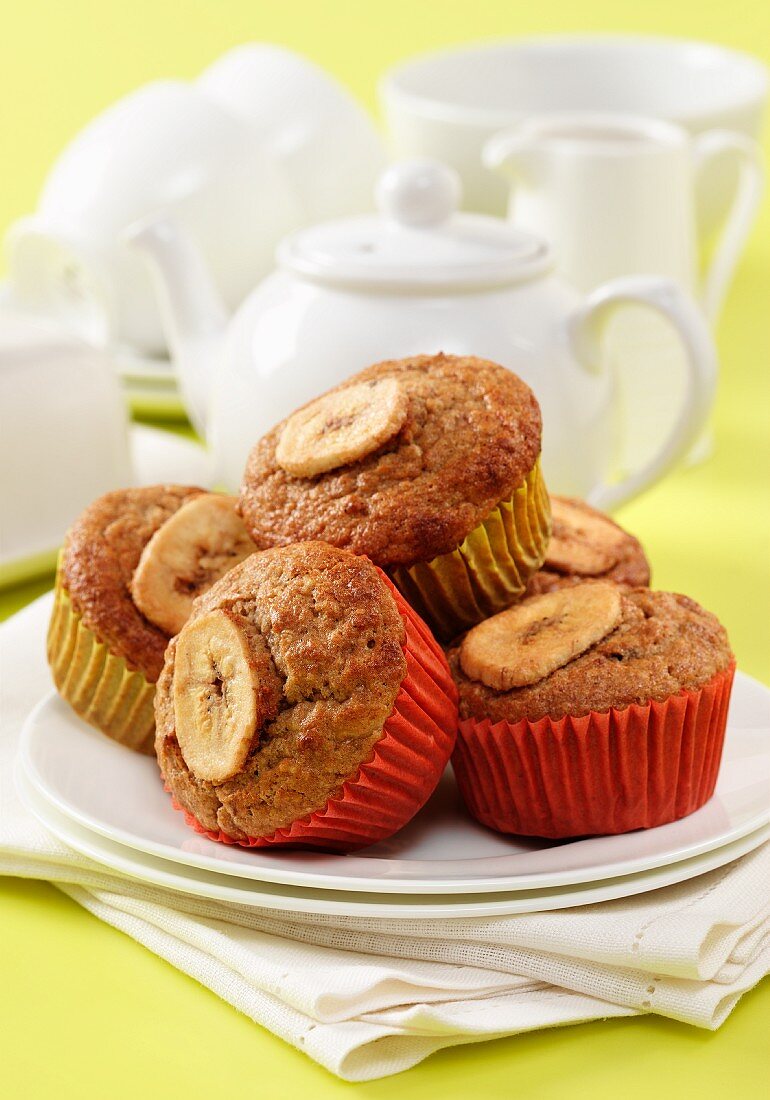 Muesli and banana muffins on a white plate with napkins and a white teaset in background sitting on a yellow surface