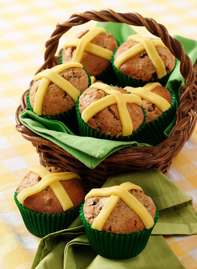 Several Easter muffins with marzipan crosses in a basket and on a napkin sitting on a gingham cloth