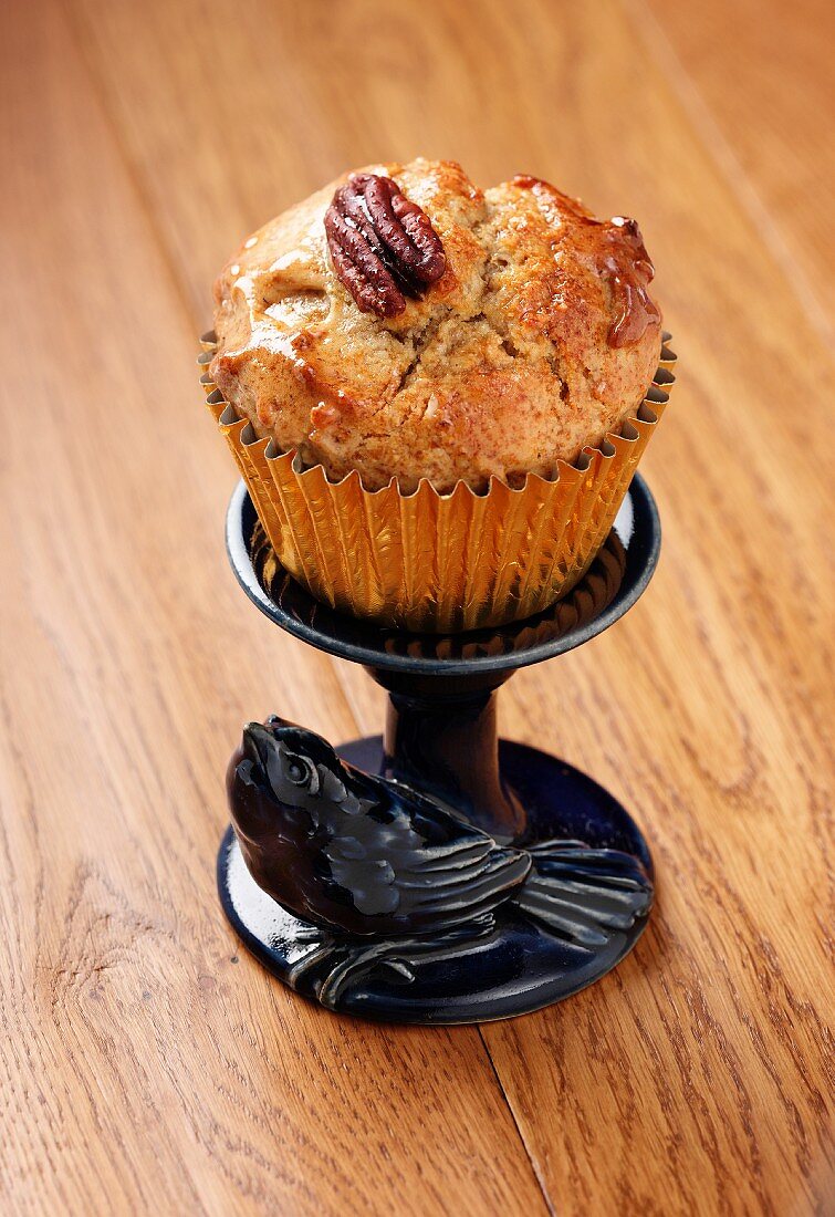 A Pecan and Maple (syrup) muffin in a gold muffin case on a small blue stand with a ceramic bird all sitting on a wooden surface