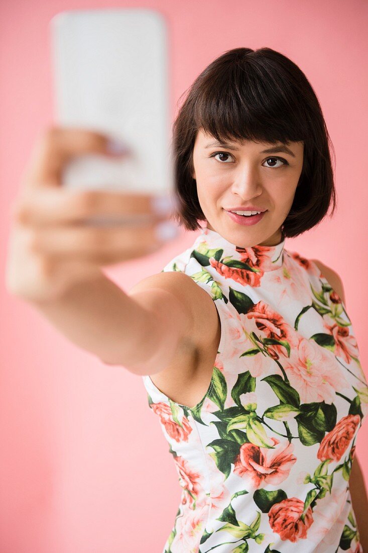A young brunette woman wearing a floral dress and taking a selfie with her smartphone