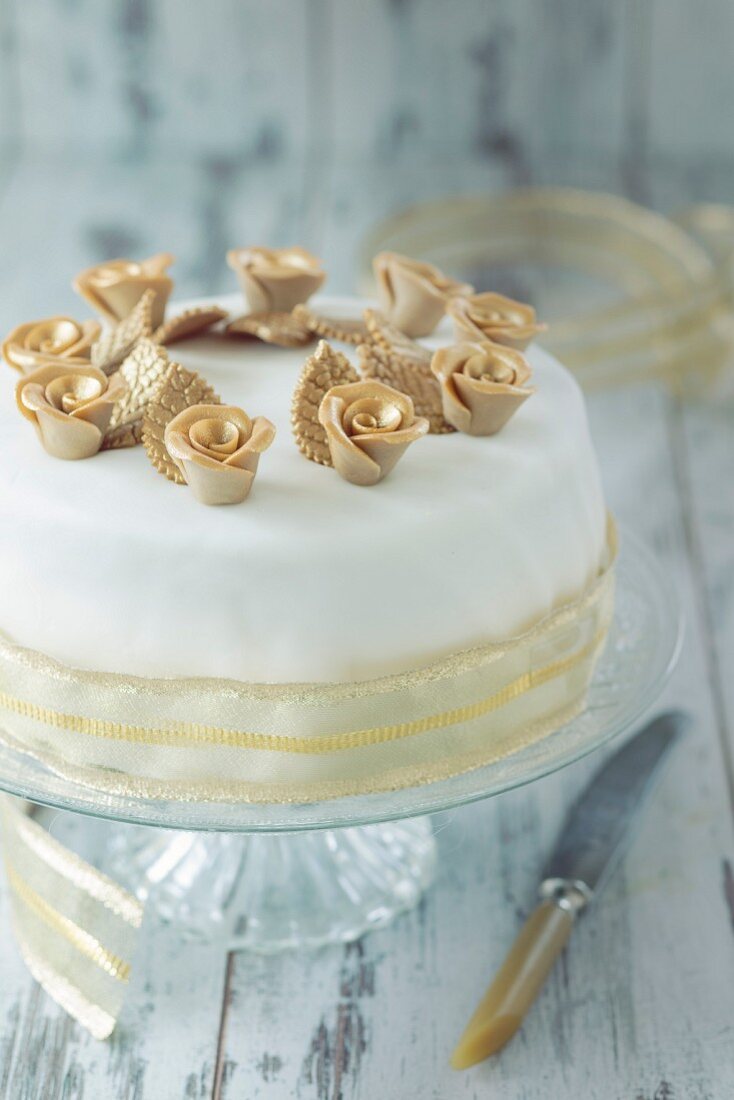 A white festive cake with gold roses and decorative ribbon