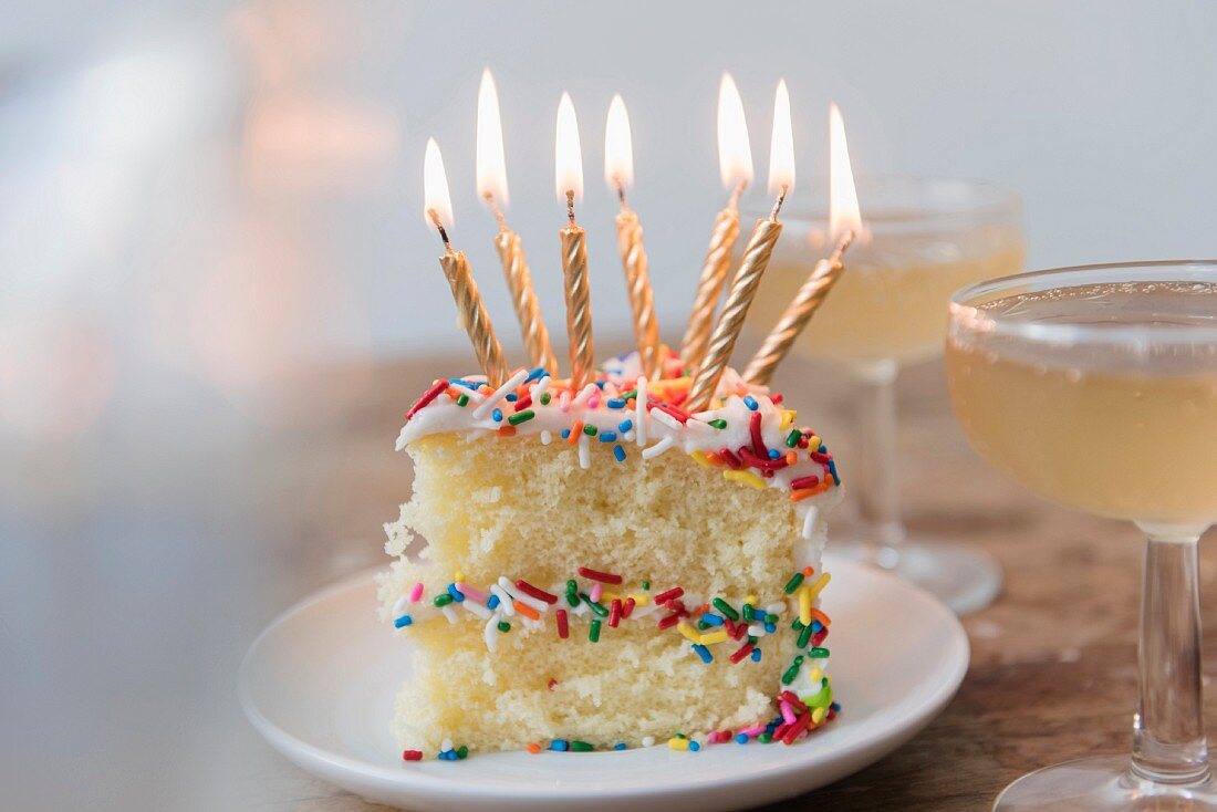 Geburtstagskuchen mit brennenden Kerzen und bunten Zuckerstreuseln neben Champagnerglas
