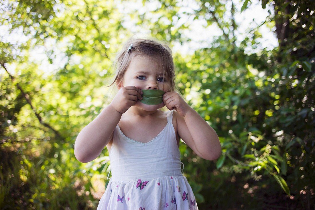 A little girl wearing a summer dress holding a leaf to her mouth