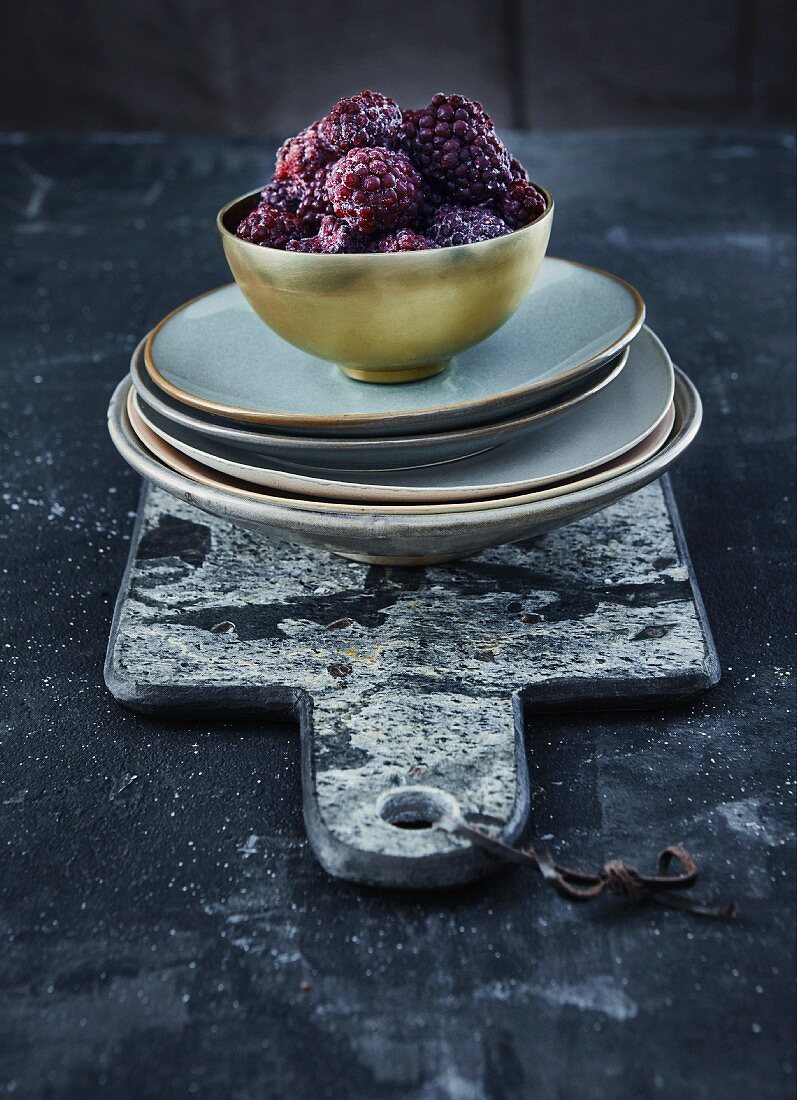 Frozen blackberries in a golden bowl on a stack of plates and a marble board