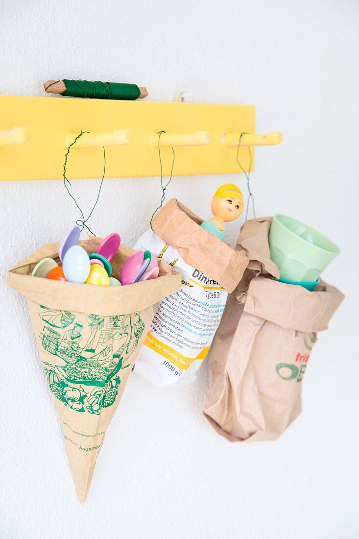 Paper bags reused to store plastic spoons and other utensils hung from yellow coat rack by florists' wire