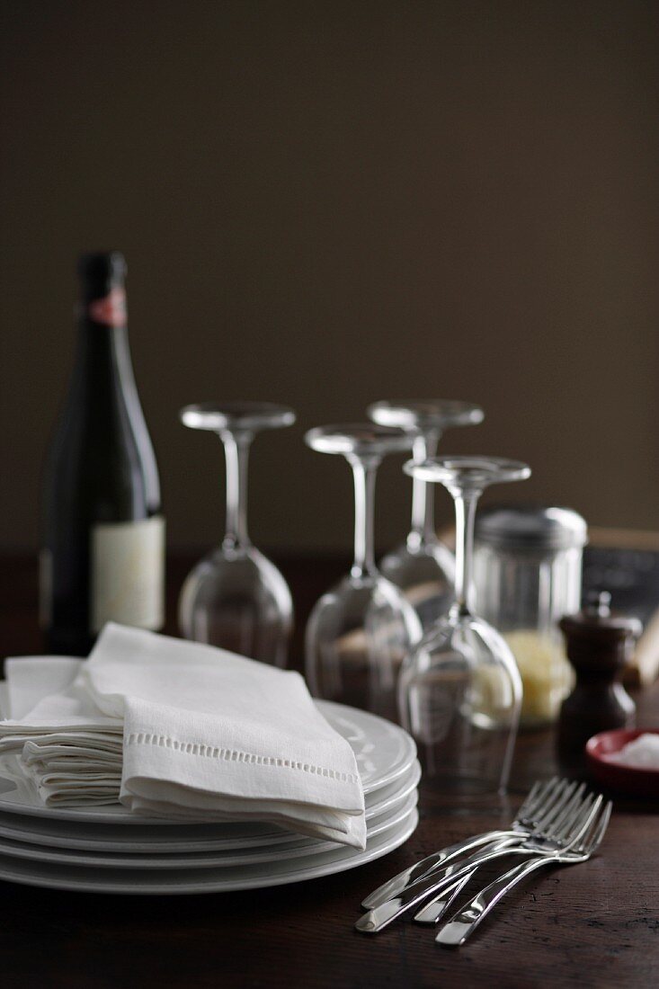 Tellerstapel, Stoffservietten, Weingläser, Weinflasche und Gewürze auf Holztisch