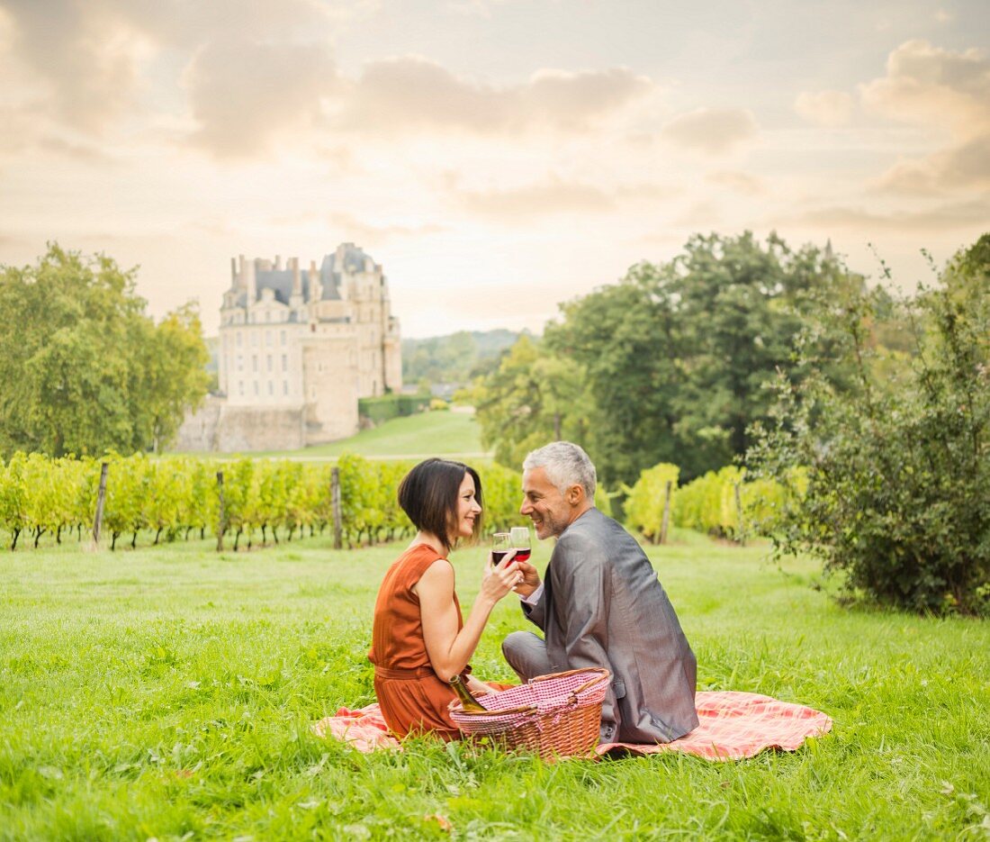 A couple drinking wine at a picnic on a meadow with a vineyard and a castle in the background