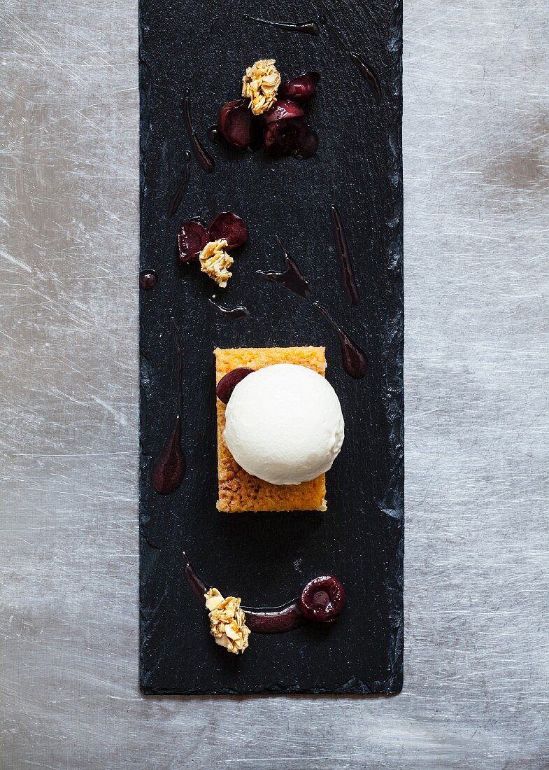 A ball of honey and rosemary parfait on a slice of carrot cake, halved cherries, cherry sauce and crispy muesli on a slate platter