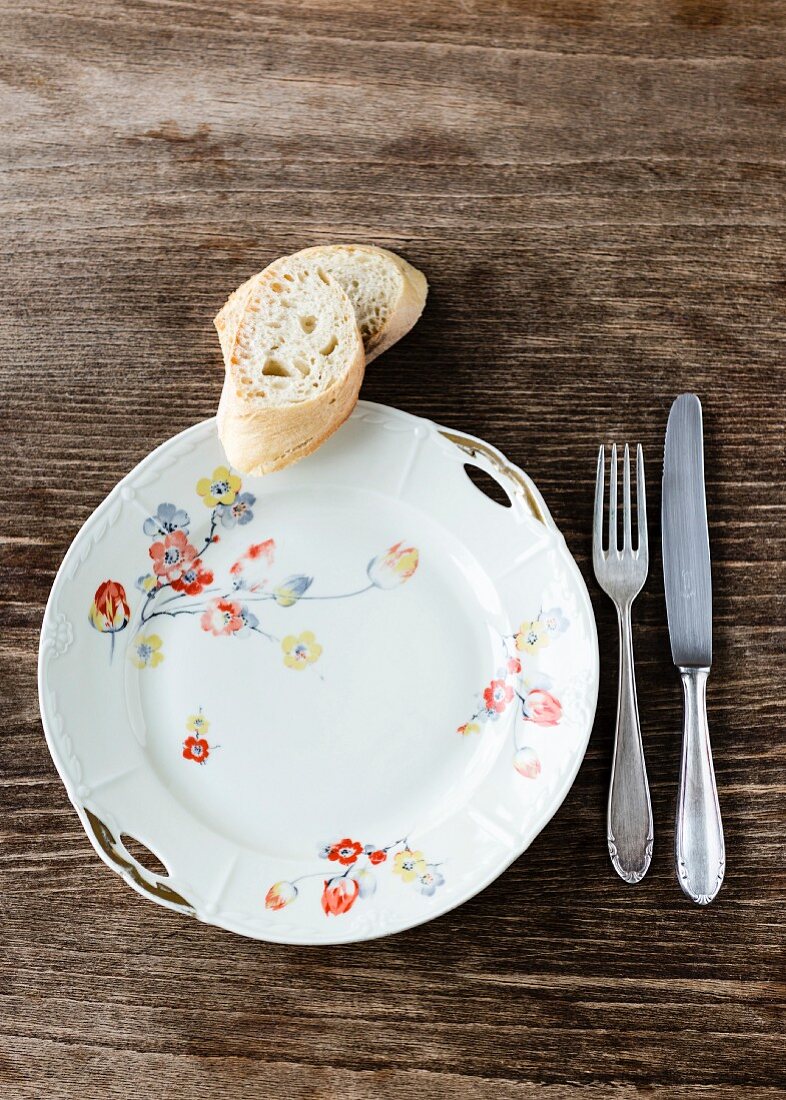 An old white porcelain plate decorated with flowers with silver cutlery and two slices of baguette on the wooden table