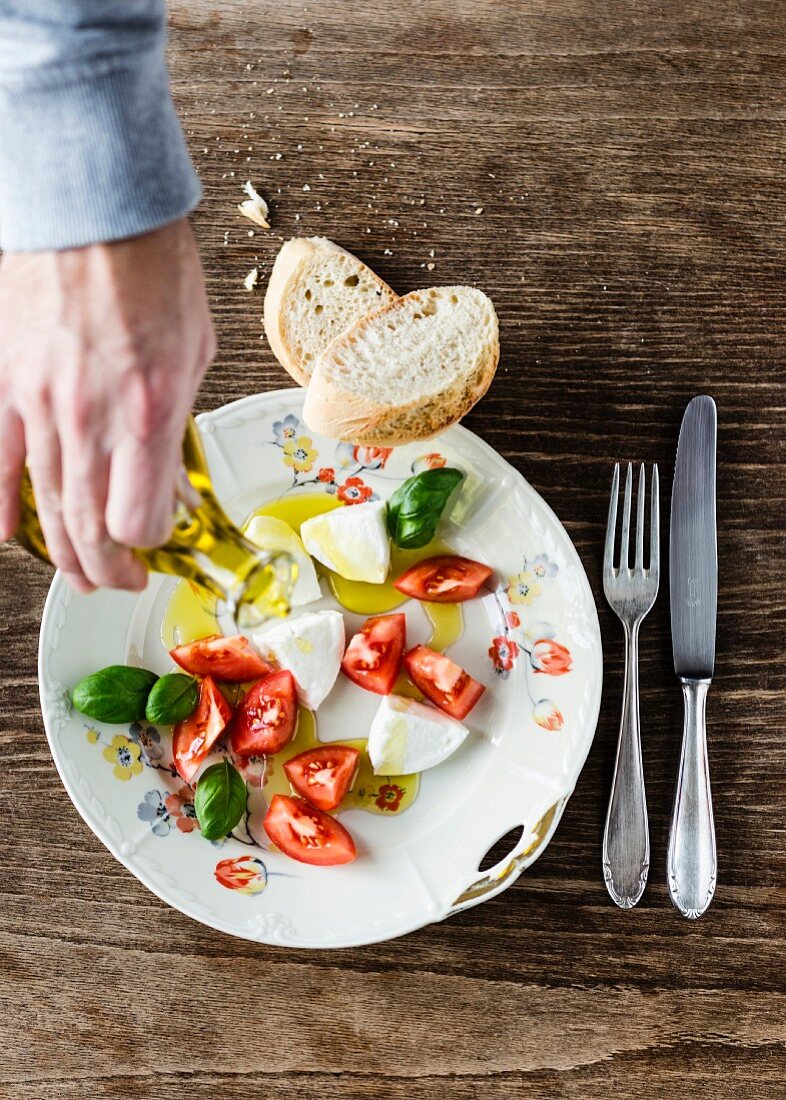 Olivenöl auf Mozzarella, Tomatenstücke, Basilikum und Baguette auf altem Porzellanteller mit Blumendekor gießen
