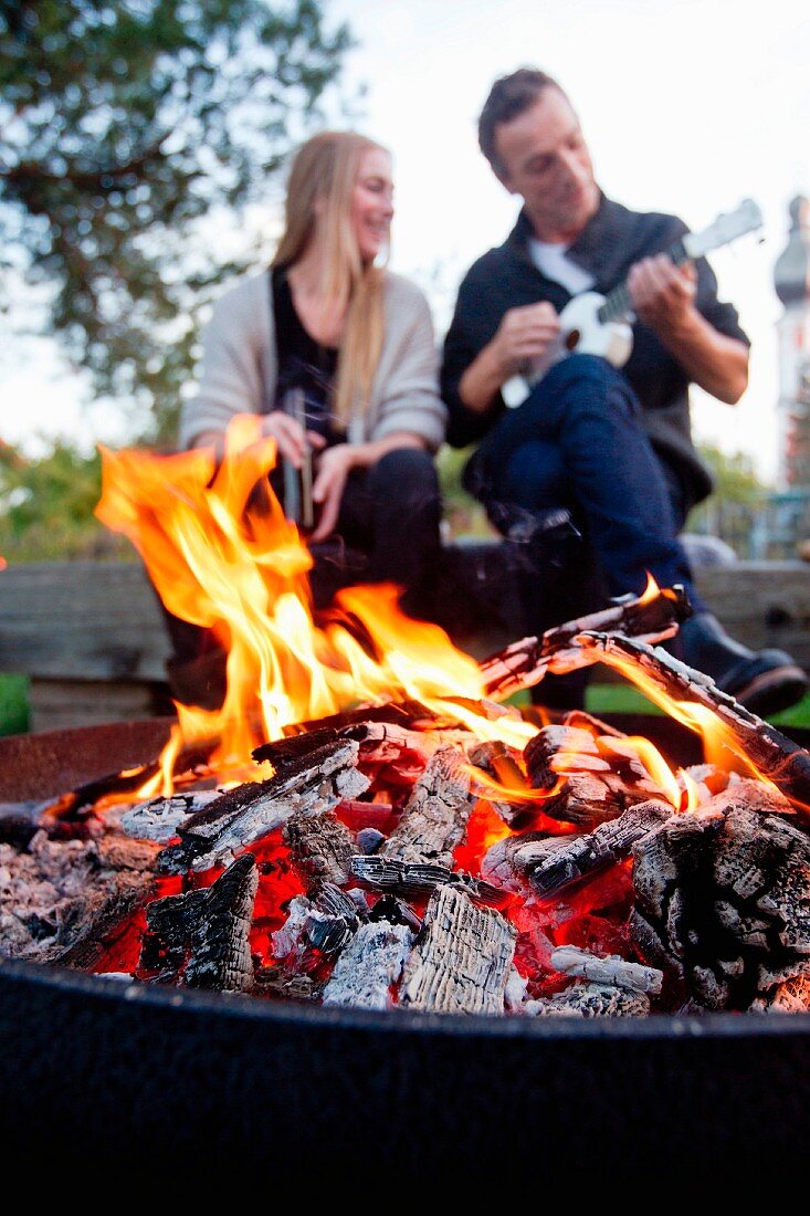 Lodernedes Feuer in einer Feuerschale, im Hintergrund ein junges Paar mit Ukulele