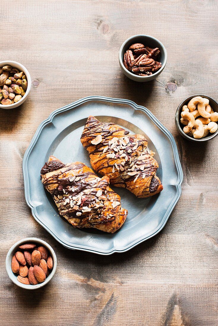 Two chocolate croissants on a plate next to various bowls of nuts