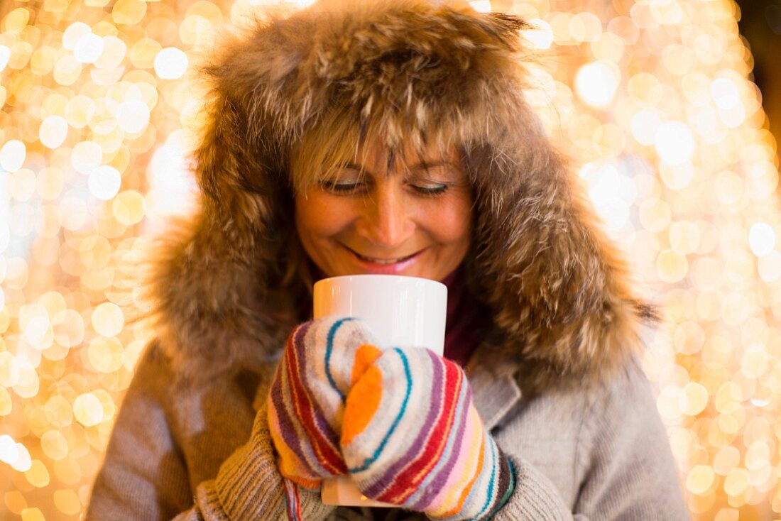 A woman wearing a furry hood and mittens holding a hot drink with Christmas lights in the background