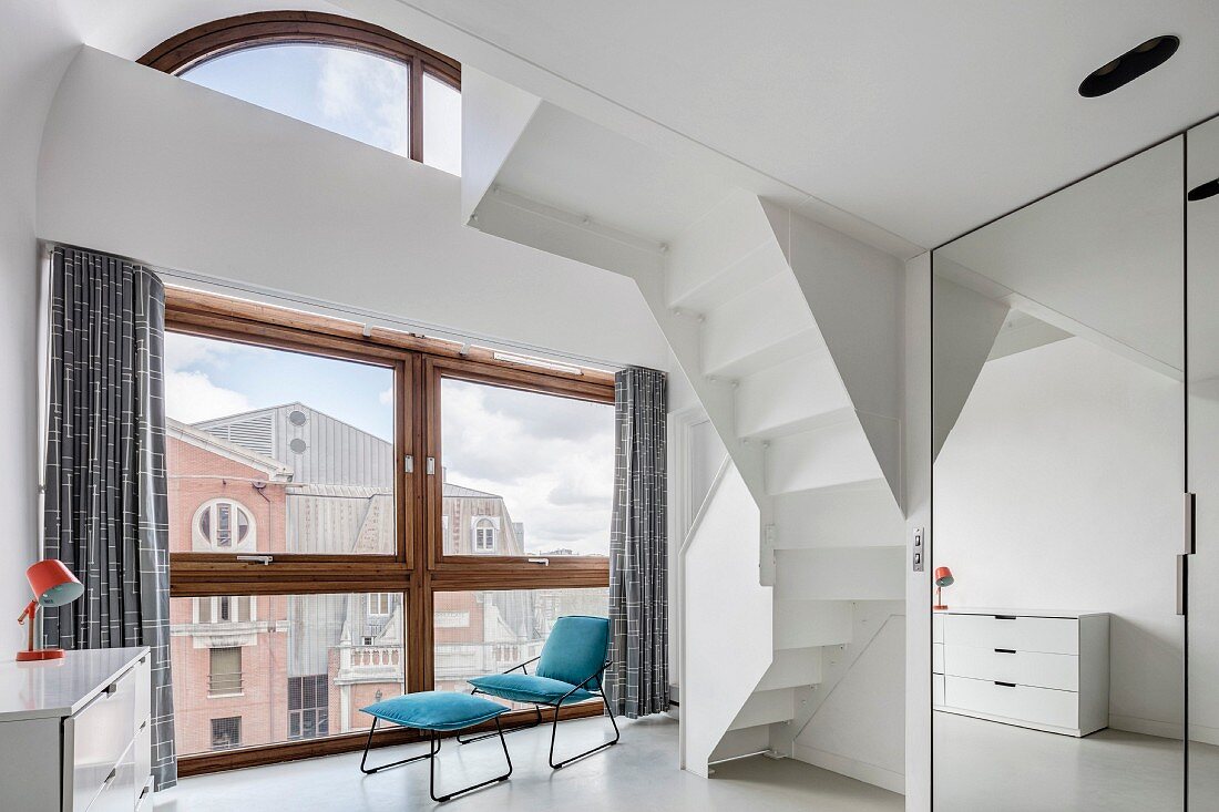 Hoher Raum mit Fensterfront und Stadtblick, Polsterstuhl mit Schemel und weiße, gewendelte Treppenuntersicht