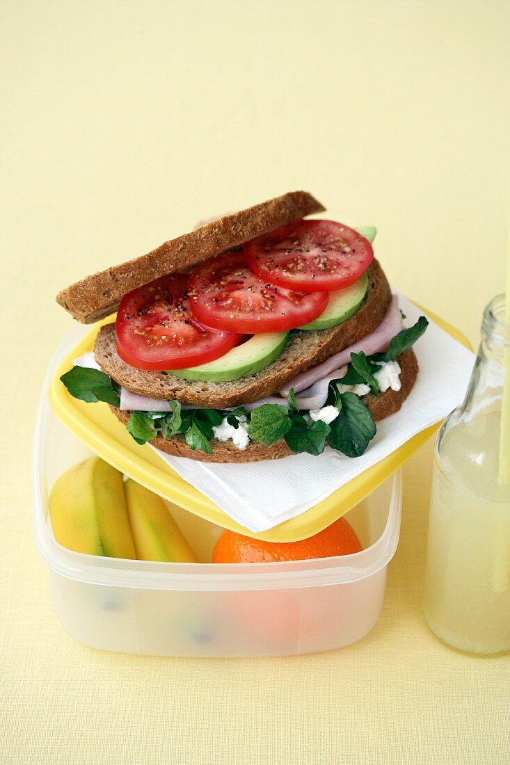 Doppeldecker-Sandwich mit Schinken, Avocado und Tomaten auf Lunchbox mit Bananen und Orange
