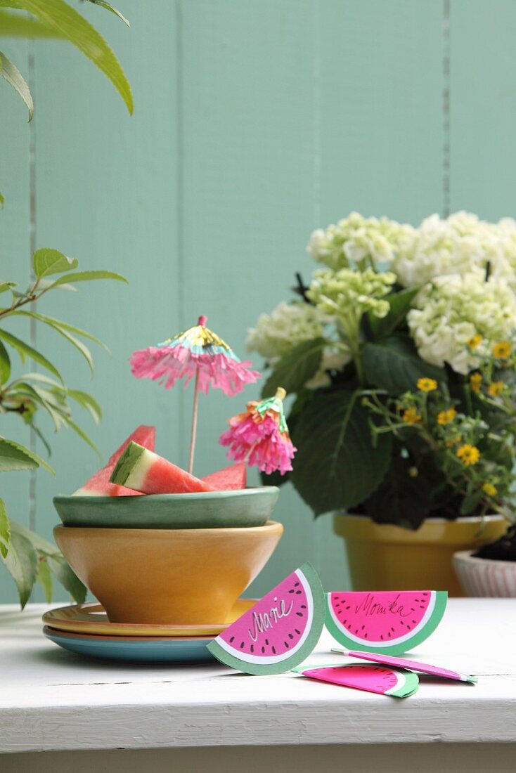Hand-made, watermelon-slice invitation cards made from folded paper next to ceramic bowls on garden table